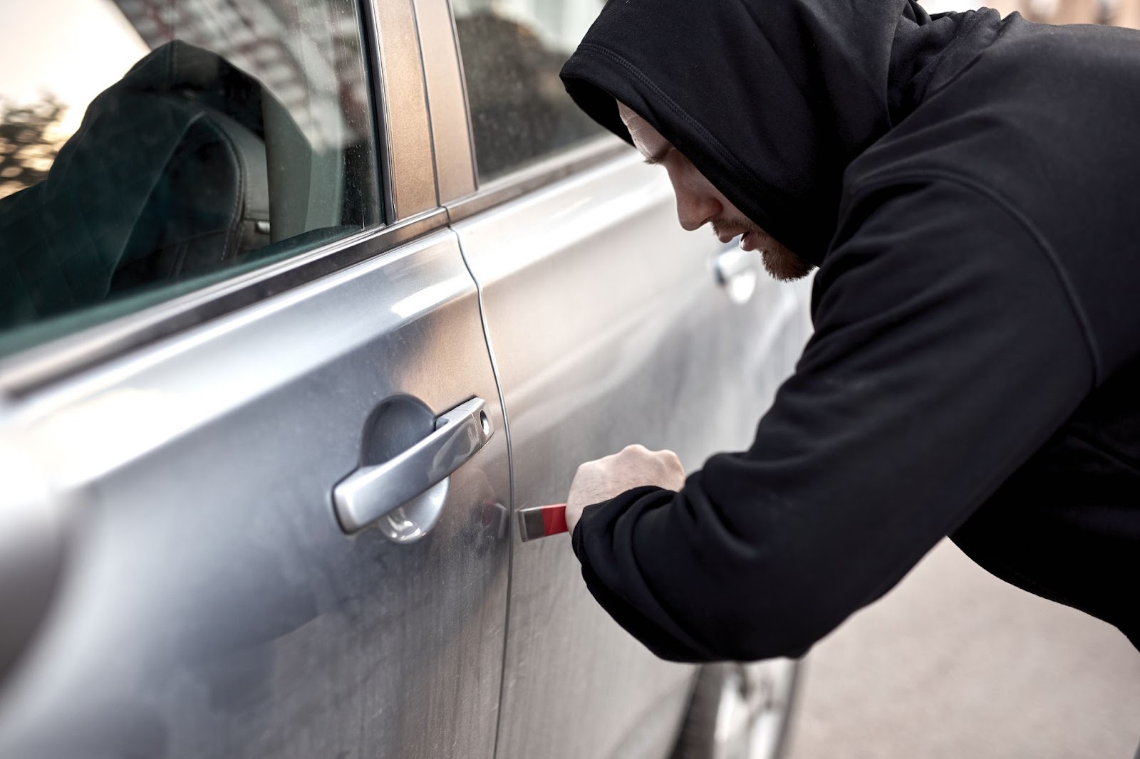 Zloděj v černé mikině s páčidlem, snažící se ukrást stříbrné auto přes přední dveře.