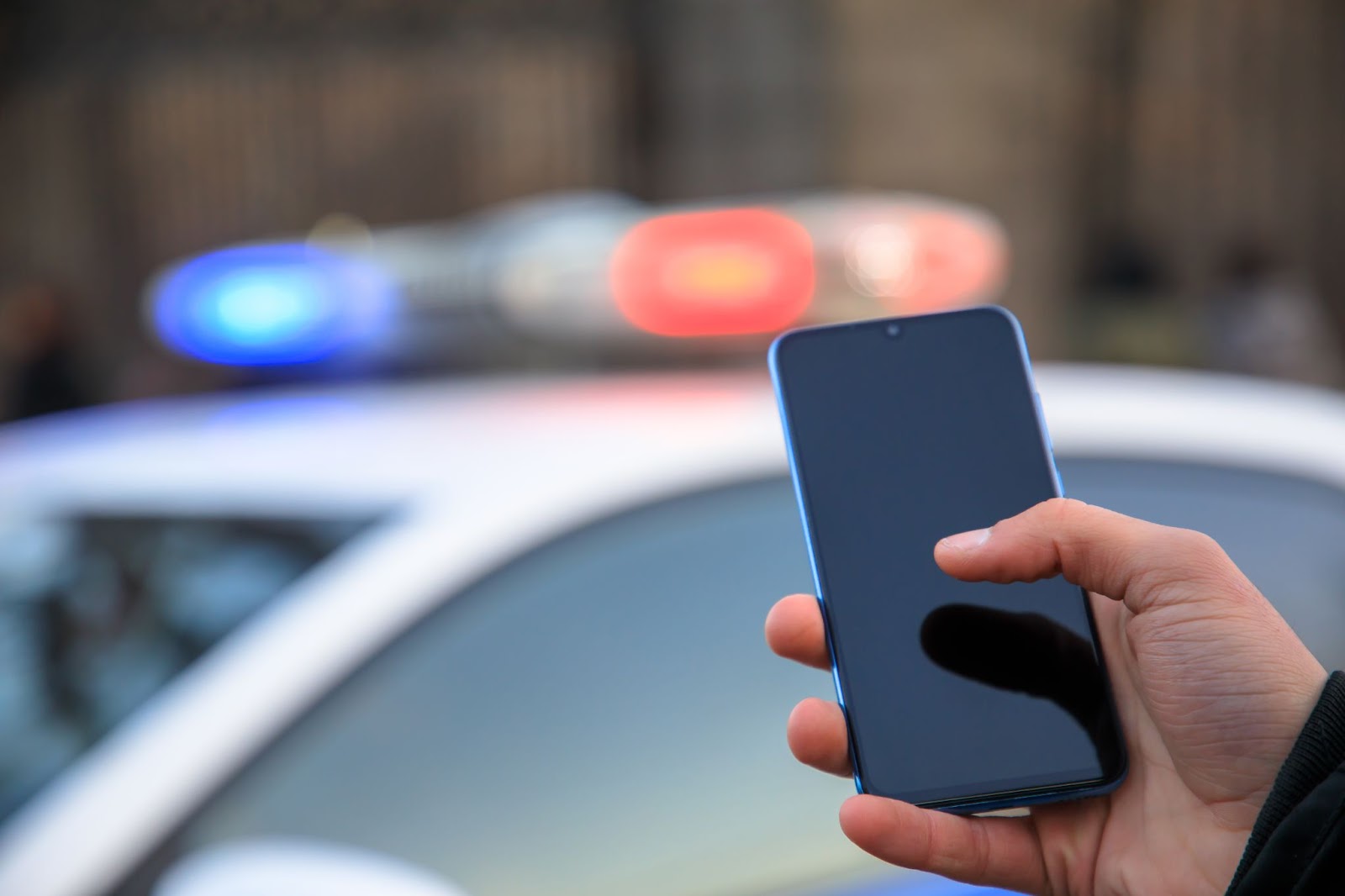 Nejdůležitější je vždy kontaktovat policii, následně pojišťovnu. | WebJET CMS Ruka držící telefon před policejním autem se zapnutými majáky.